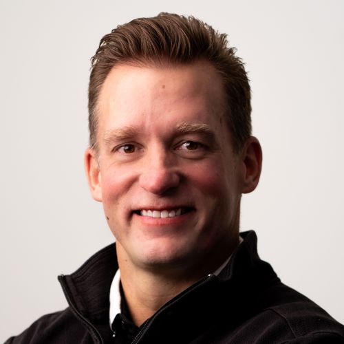 Head-and-shoulders portrait of a smiling man in a black zip-up sweater, studio lighting; professional headshot.