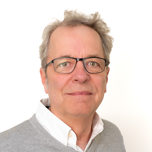 Portrait of a middle-aged man with grey hair and glasses, wearing a white shirt and light grey sweater, looking at the camera.