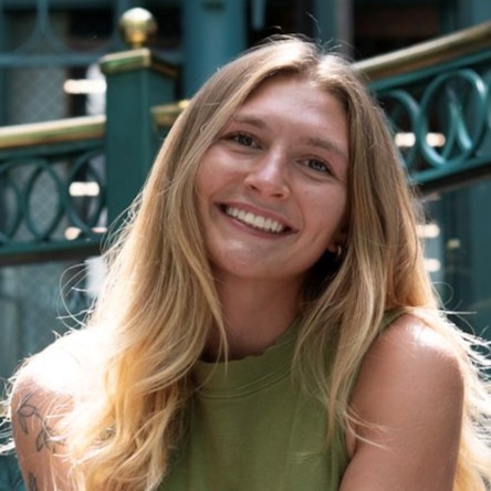 Smiling woman with long blonde hair in a green sleeveless top, outdoors near a railing.
