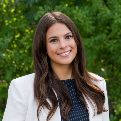 Smiling woman with long brown hair wearing a white blazer, outdoors with green foliage behind her.