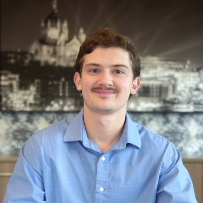 Young man in a blue shirt smiling at the camera, seated indoors with a grayscale cityscape backdrop in the background.