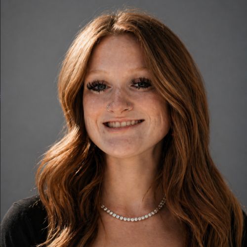 Portrait of a smiling woman with long red hair, wearing a pearl necklace against a gray backdrop.